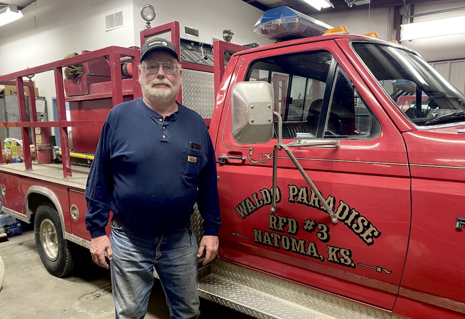 Caption: Keith Koelling is 62 and still fighting fires.  He says he worked 40 straight hours on a recent fire, only to find out he had COVID-19 at the time.  He stands next to a 26-year-old fire engine.