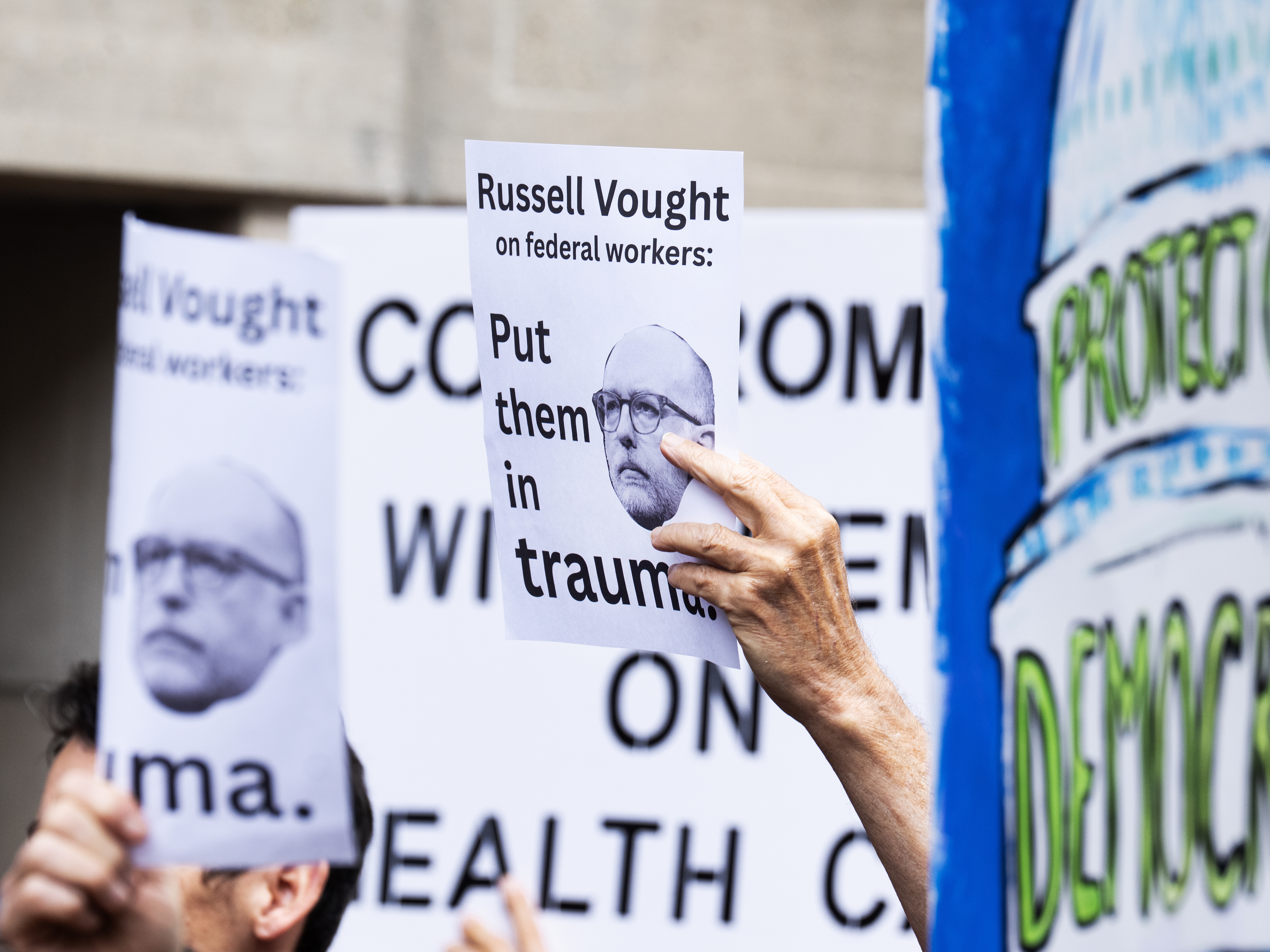 caption: Signs with the image of Office of Management and Budget Director Russell Vought are seen during a news conference Congressional Democrats held to protest the Trump administration's mass firing of federal employees in Washington, D.C., on Tuesday, Oct. 14.