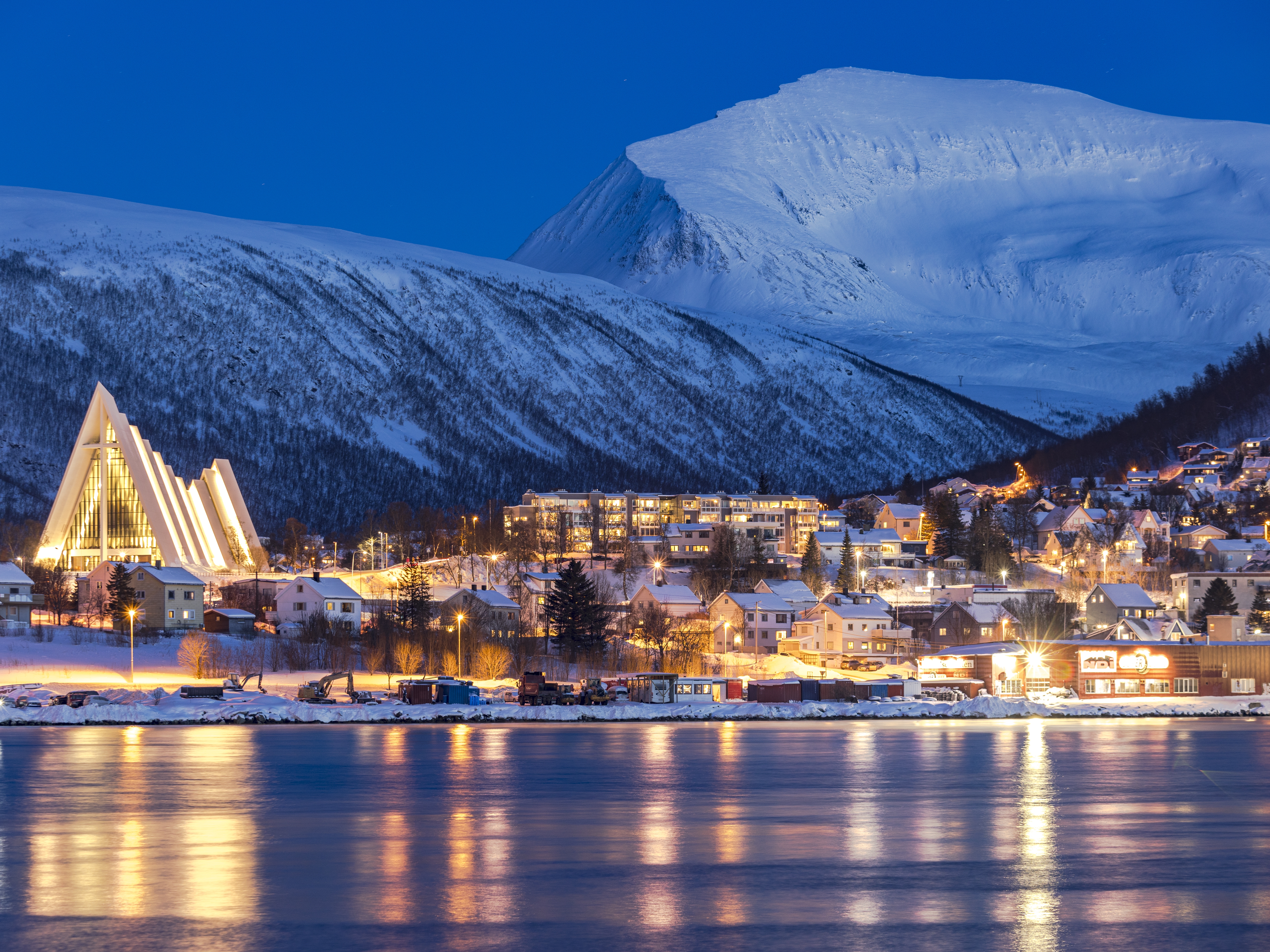 caption: Dusk falls over snowy mountains that overlook the Arctic Cathedral and the houses mirrored in the icy fjord in Tromsø, Norway. Although this city is north of the Arctic Circle and experiences polar night for two months out of the year, its residents tend to see winter as a time of opportunity.