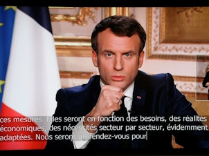 caption: French President Emmanuel Macron is seen on a television screen as he speaks during an address to the nation on the outbreak of COVID-19, caused by the novel coronavirus, on Monday.