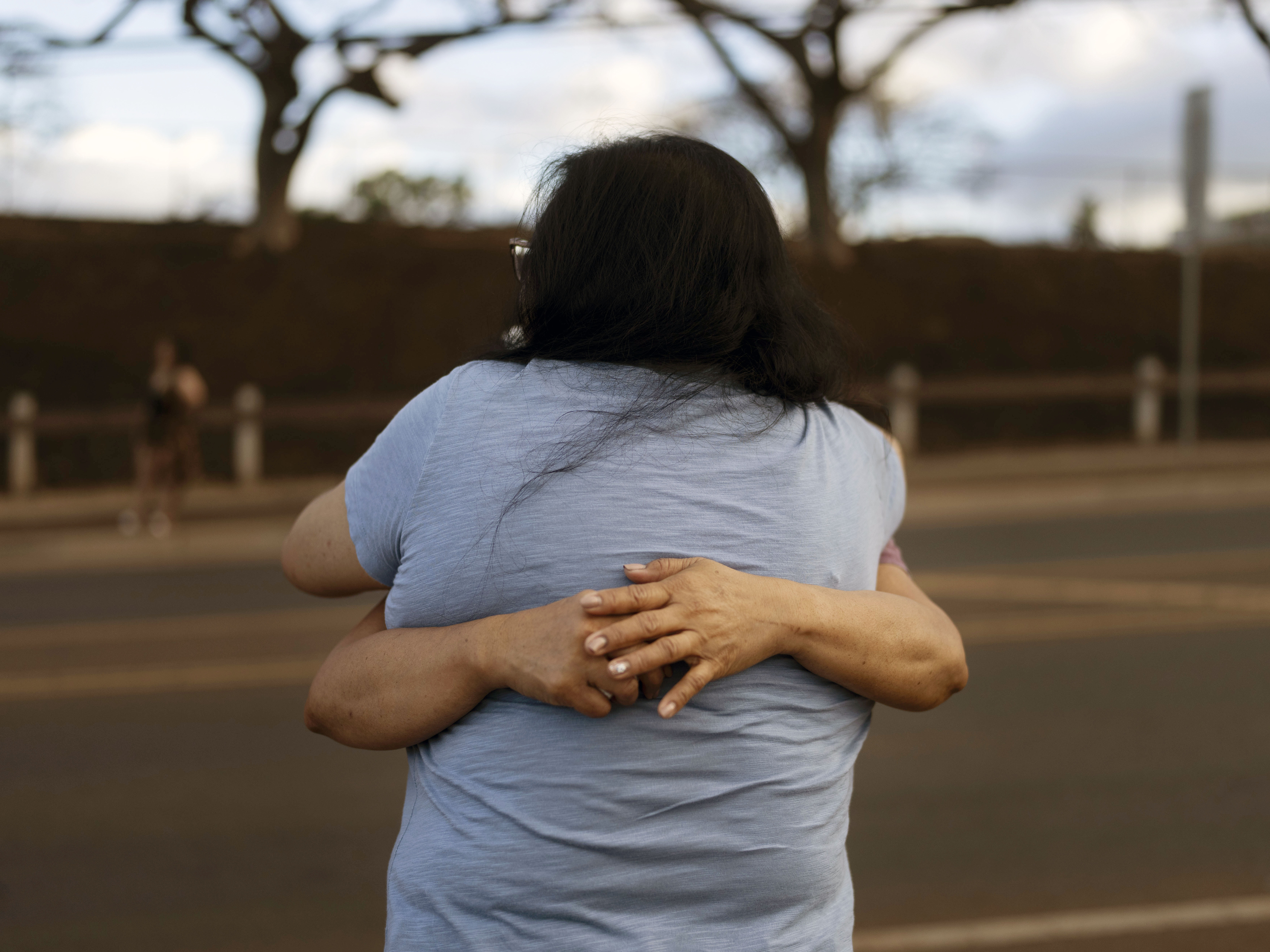 caption: Carelle Calvan in blue T-shirt, hugs Nora Bulosan as they gather near a checkpoint in hopes to get access to their home destroyed in recent wildfires in Lahaina, Hawaii, Wednesday, Aug. 16, 2023.