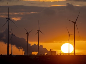 caption: Steam rises from the coal-fired power plant with wind turbines nearby in Niederaussem, Germany, as the sun rises on Nov. 2, 2022.