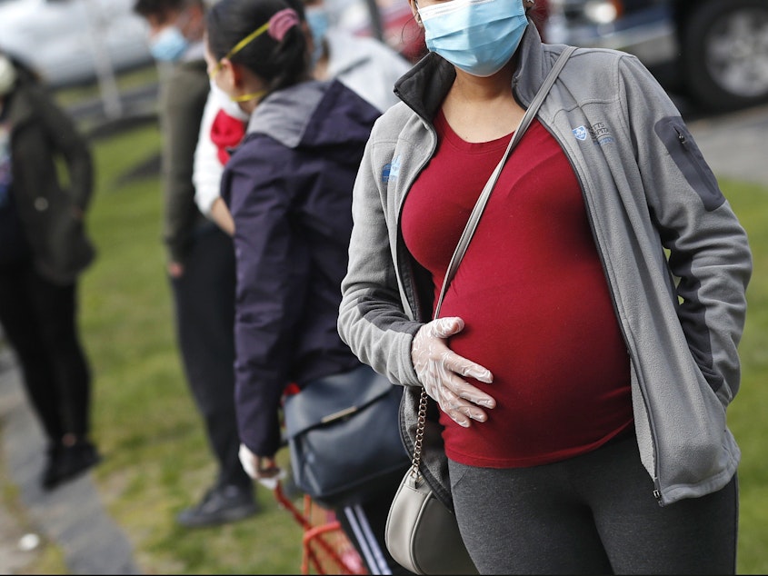 caption: A pregnant woman wearing a face mask and gloves holds her belly as she waits in line for groceries at St. Mary's Church in Waltham, Mass., in May. The Centers for Disease Control and Prevention on Wednesday again urged all pregnant women to get the COVID-19 vaccine.