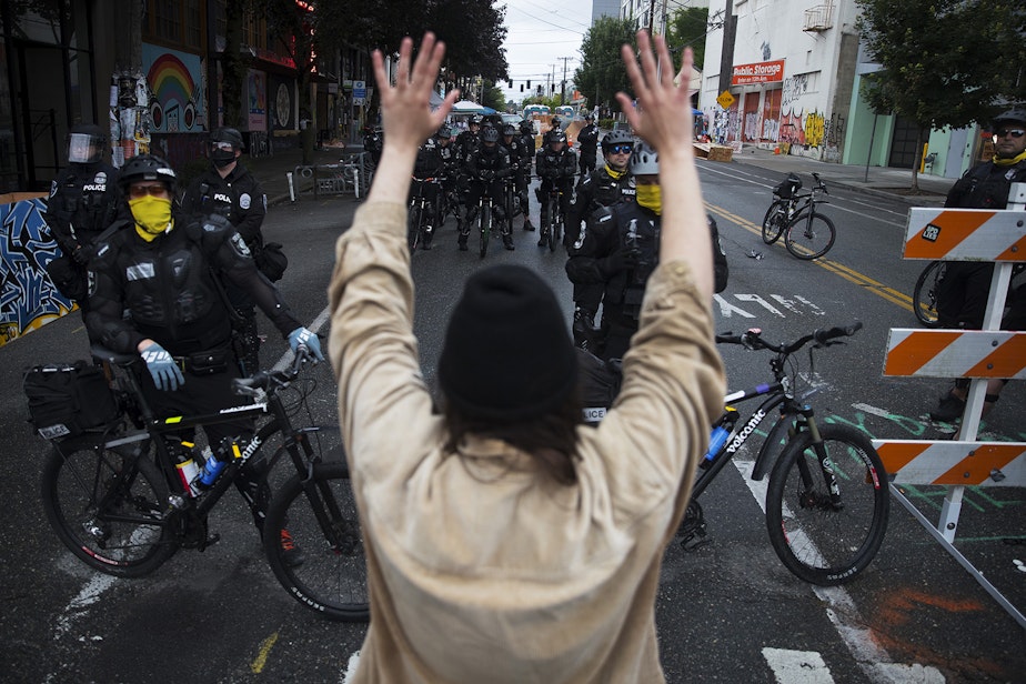 caption: A protesters raises their hands in the air while standing at a police line outside of the East Precinct building after the Capitol Hill Organized Protest zone was cleared by Seattle Police Department officers early Wednesday morning, July 1, 2020, at the intersection of 12th Avenue and in East Pike Street in Seattle. 
