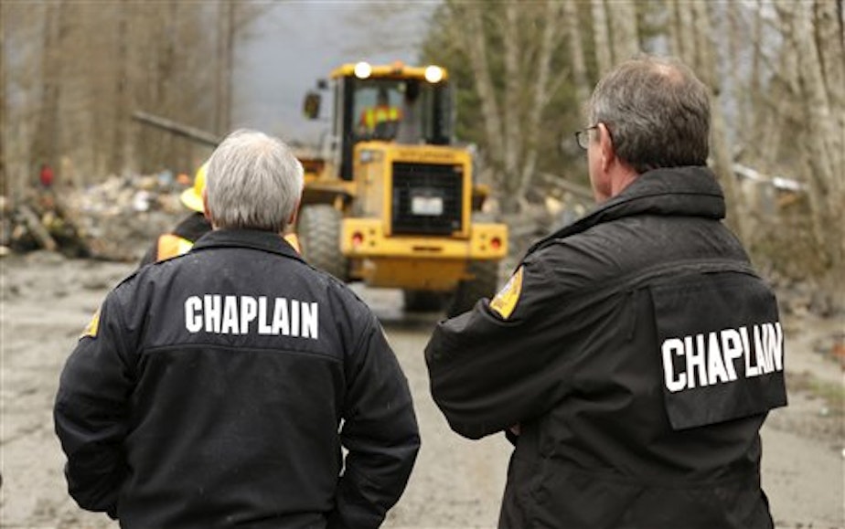 caption: Washington State Patrol chaplain Mike Neil, right, looks on with a colleague as workers using heavy equipment work to clear debris from the Oso mudslide.