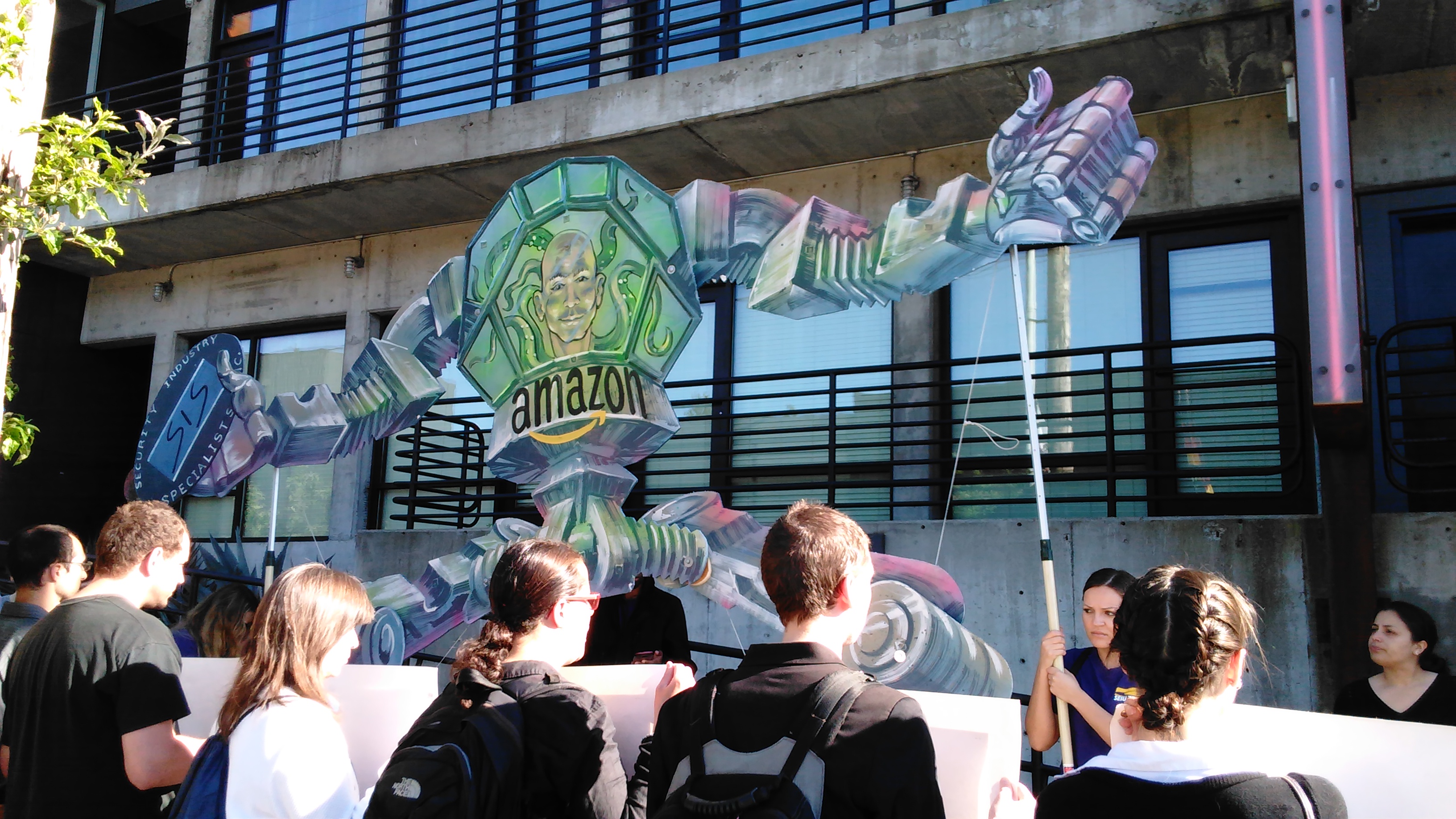 caption: Protesters outside Amazon's annual shareholder's meeting hold up a RoboBezos sign