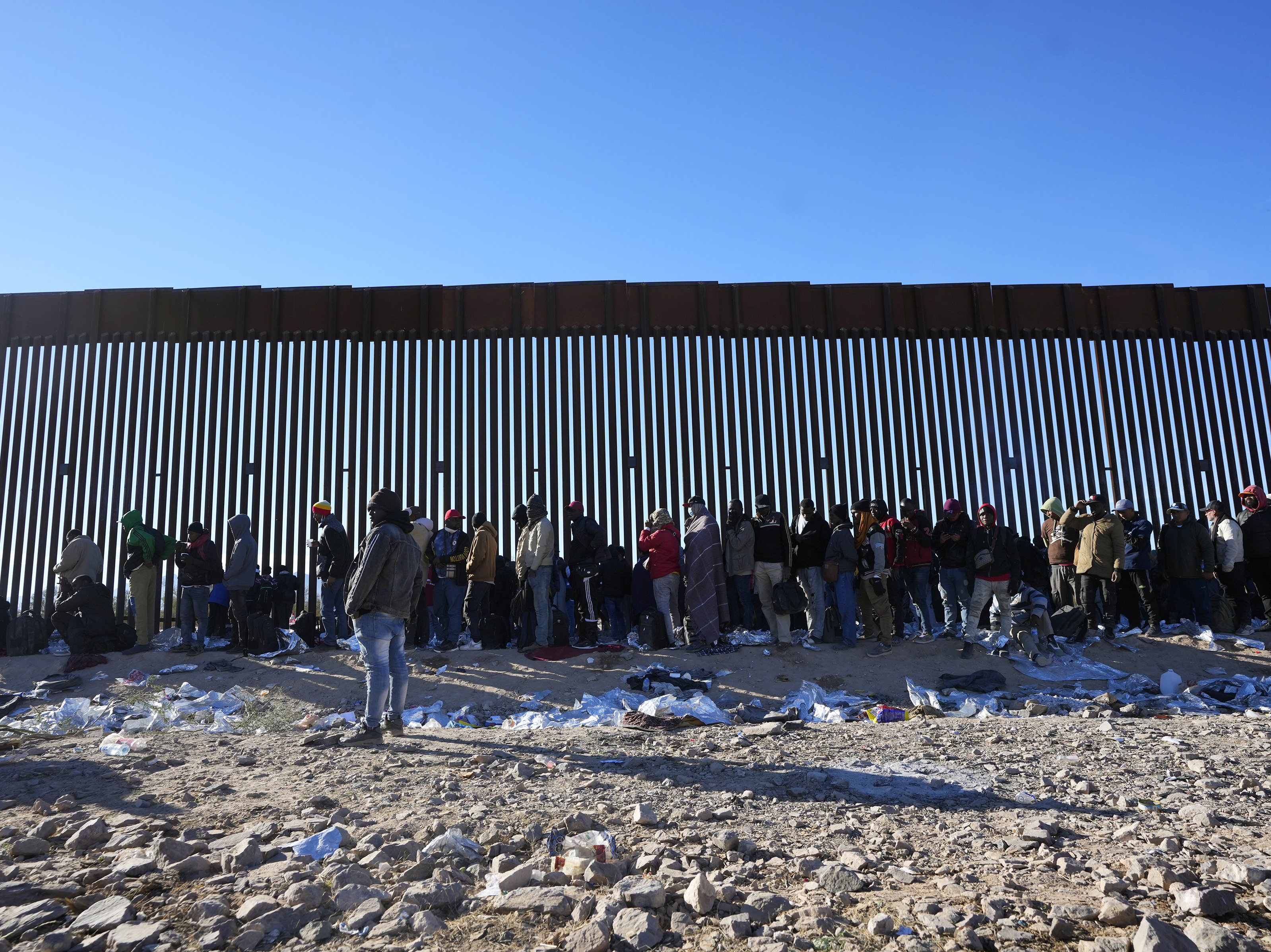 caption: Hundreds of migrants gather along the border wall Tuesday, Dec. 5, 2023, in Lukeville, Ariz. The U.S. Border Patrol says it is overwhelmed by a shift in human smuggling routes.