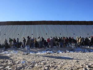 caption: Hundreds of migrants gather along the border wall Tuesday, Dec. 5, 2023, in Lukeville, Ariz. The U.S. Border Patrol says it is overwhelmed by a shift in human smuggling routes.