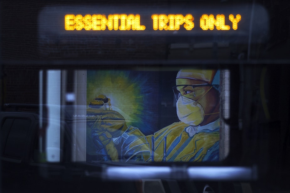 caption: A mural of a healthcare worker wearing personal protective equipment is shown through the window of a passing King County Metro bus on April 28, 2020, along South Main Street in Seattle. As Seattle businesses, coffee shops and restaurants shut down amid the pandemic, plywood replaced their many entrances and windows. Artists transformed that plywood into murals bearing messages of hope and resilience.