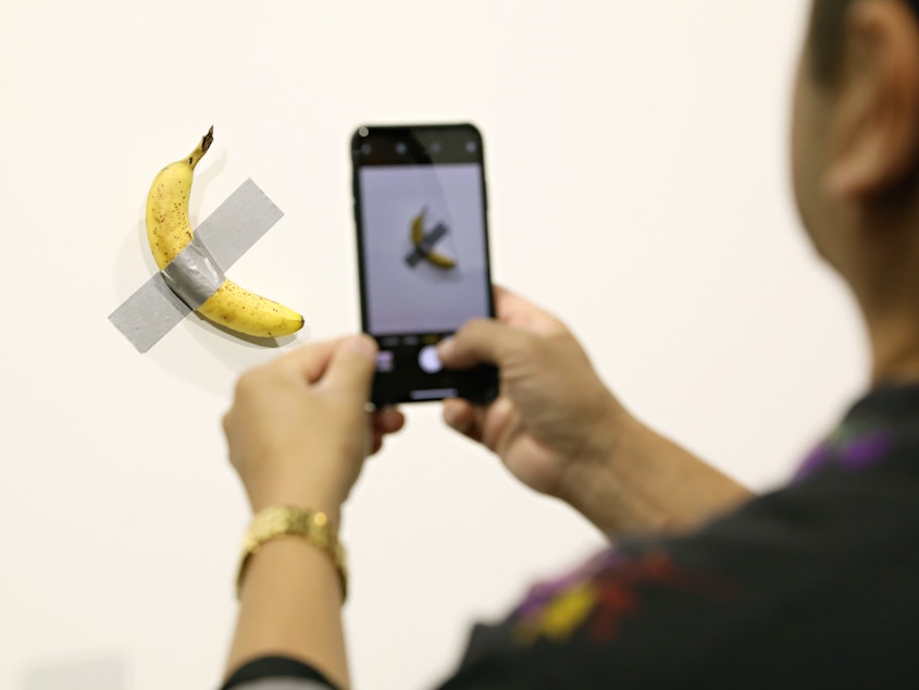 caption: People view Maurizio Cattelan's "Comedian" on view at Art Basel Miami 2019 Friday. Two of the three editions of the piece, which feature a banana duct-taped to a wall, have reportedly sold for $120,000.