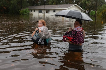 caption: Makatla Ritchter and her mother, Keiphra Line, wade through flood waters after evacuating their home in Tarpon Springs, Florida. Flood waters from Hurricane Idalia inundated it on August 30, 2023. Climate change is making storm surge and intense rainfall during hurricanes like Idalia more dangerous.
