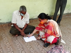 caption: Prema Thakur, an official for the Champawat district in India, teaches Pratap Singh Bora, a 56-year-old migrant laborer from Nepal, how to write his name in Hindi.
