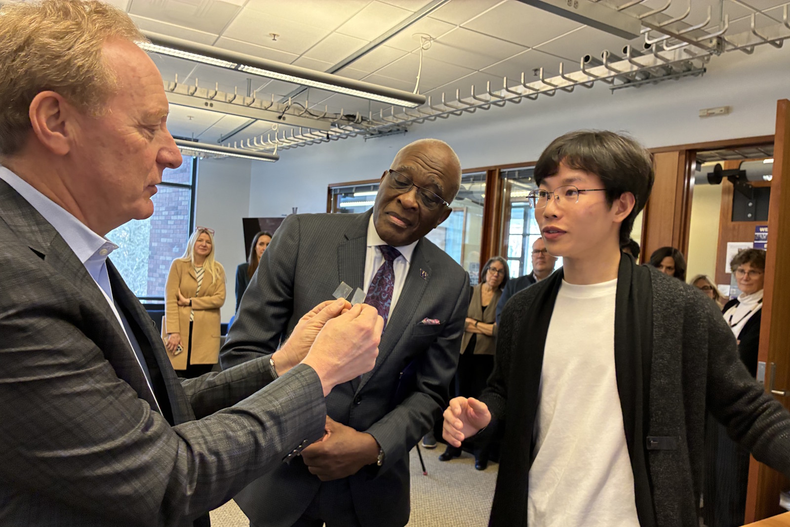 caption: Microsoft Vice Chair and President Brad Smith inspects Univeristy of Washington student Zhihan Zhang's microchip materials made from recycled older chips as UW President Robert Jones looks on. Zhang has another project that uses AI to determine how many carbon emissions AI produces.