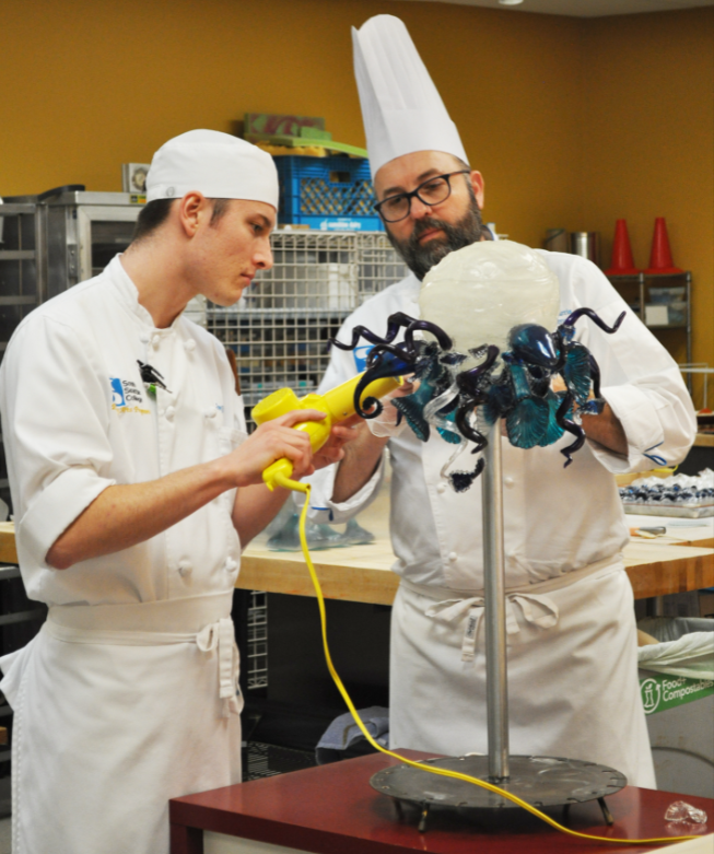 caption: Chef Christopher Harris (right) with student Joey Bale work on a replica of Dale Chihuly's glass chandelier for the artist's birthday cake.