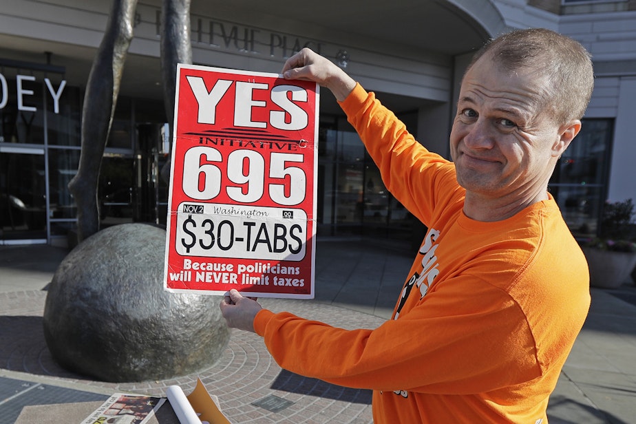 caption: Anti-tax activist Tim Eyman poses for a photo with a sign from his original 1999 campaign for $30 car registration tabs, Tuesday, Nov. 5, 2019, as he waved a sign for his current Initiative 976 on election day in Bellevue, Wash. The measure would again cut most car tabs to $30 in Washington state, if passed by voters, and would leave state and local governments scrambling to pay for road paving and other transportation projects. 