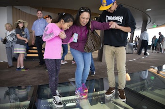 caption: Samantha Ramirez, 9, left, and Benjamin Ramirez, 16, right, help their mother, Sheila Ramirez, center, to step onto the rotating glass floor at the Space Needle on Friday, August 3, 2018, in Seattle. 