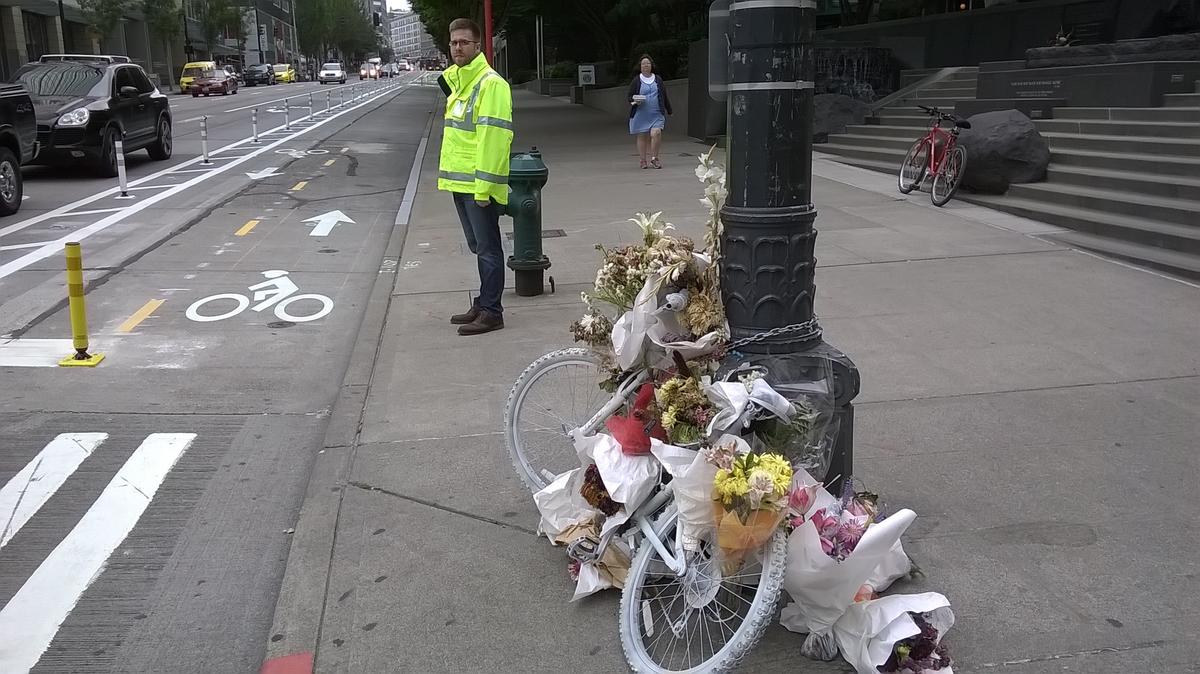 caption: Ross McFarland of the Seattle Department of Transportation prepares to flag confused drivers on the opening day of downtown Seattle's new protected bike lane. In the foreground, a memorial to Sher Kung, a cyclist who died here about two weeks ago.
