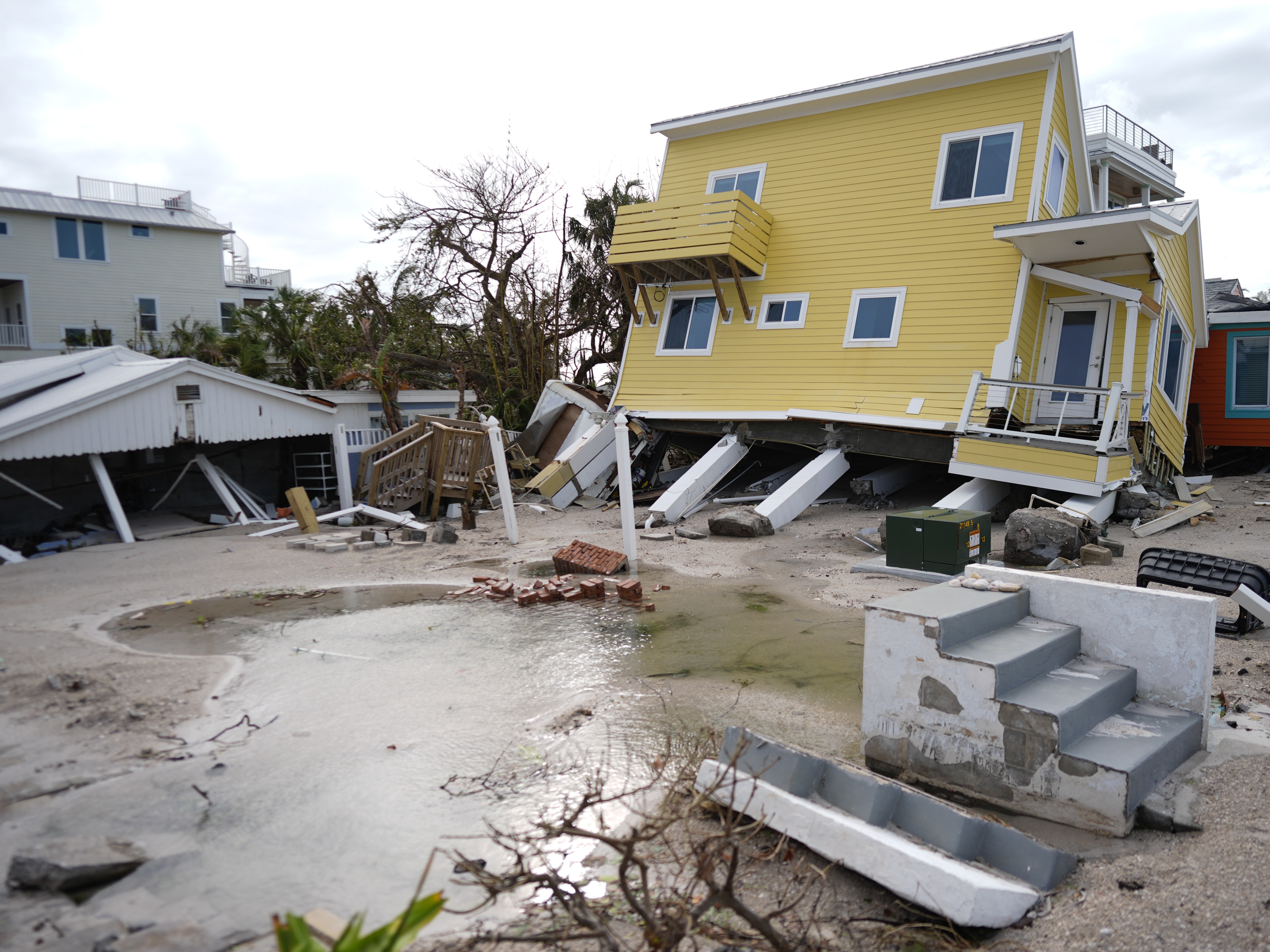 caption: A house, center, lies toppled off its stilts after the passage of Hurricane Milton, alongside an empty lot where a home was swept away by Hurricane Helene, in Bradenton Beach on Anna Maria Island, Fla.