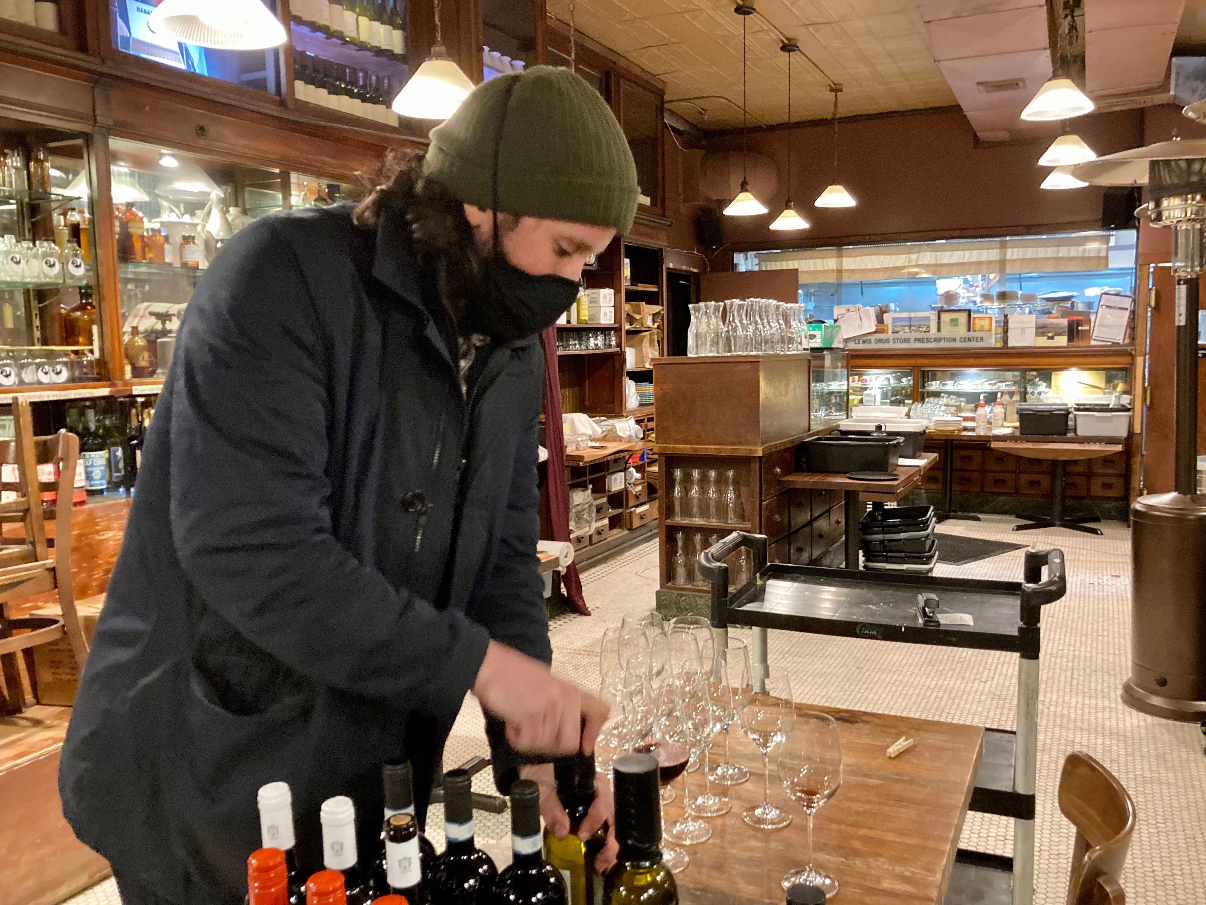 caption: Michael Schall pours wine in Vini E Ollie's dining room which has been turned into a storage space during the pandemic.