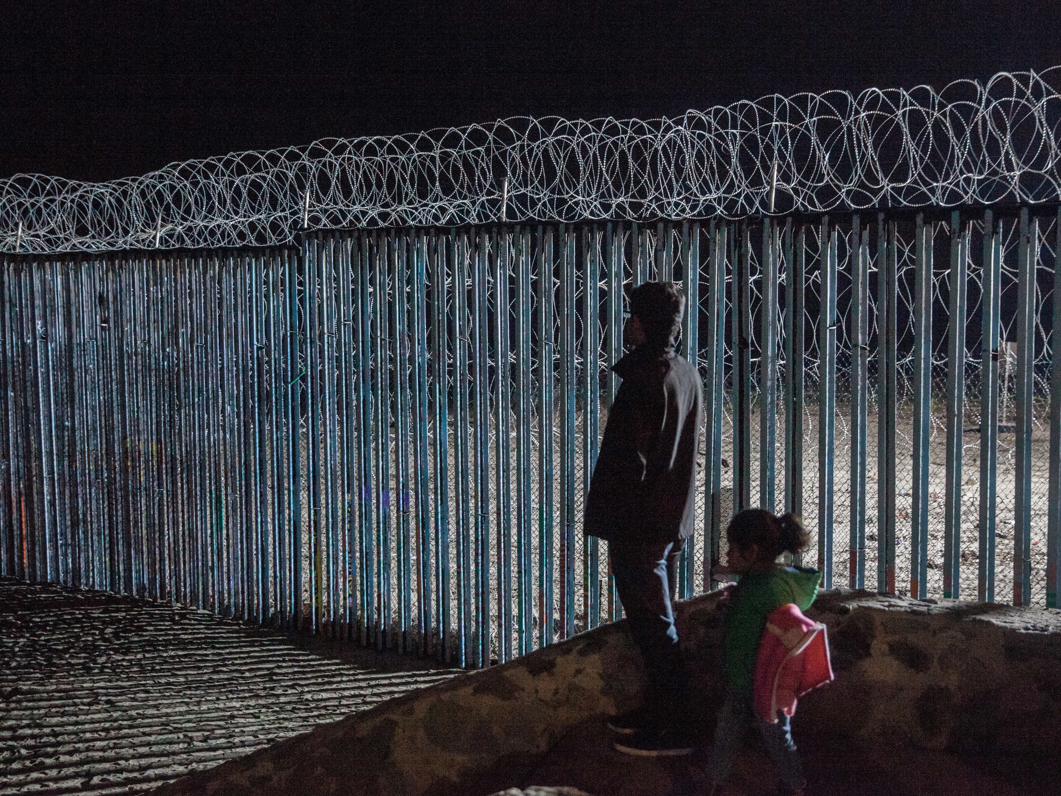 caption: A young man and a little girl look at the border fence from Playas de Tijuana, Mexico. Photojournalist Ariana Drehsler has been covering the caravan of migrants for weeks. In December, Customs and Border Protection agents began pulling her over for questioning each time she crossed back into the U.S.