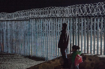 caption: A young man and a little girl look at the border fence from Playas de Tijuana, Mexico. Photojournalist Ariana Drehsler has been covering the caravan of migrants for weeks. In December, Customs and Border Protection agents began pulling her over for questioning each time she crossed back into the U.S.