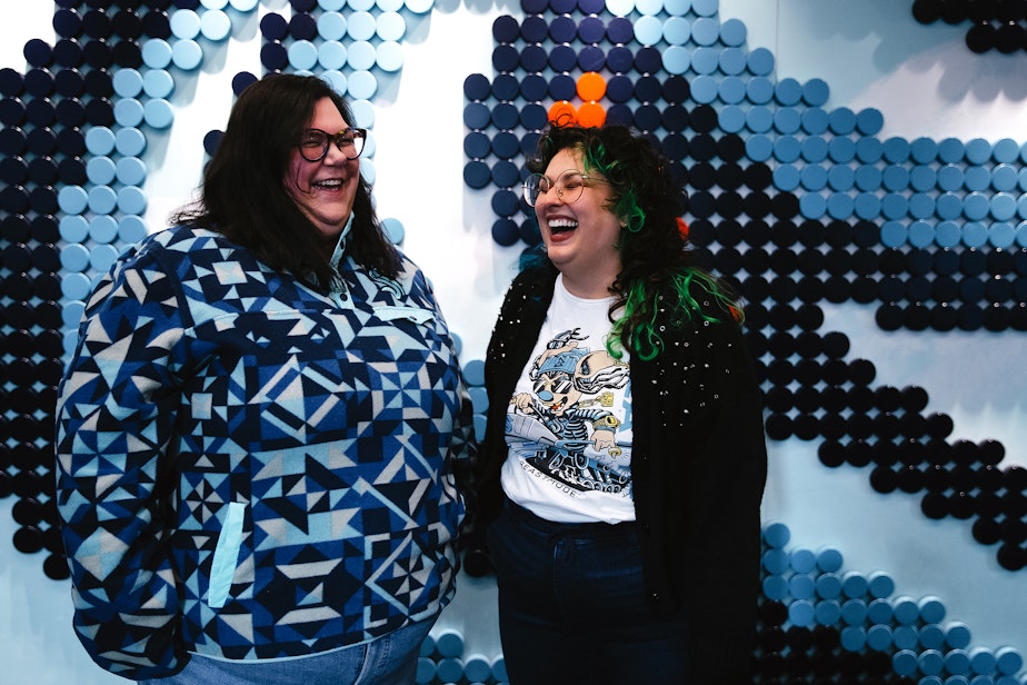 caption: Ashley Hufford, left, and Gina Rome, right, laugh together while being portrayed in front of the ‘puck wall’ on Tuesday, April 29, 2025, at the Kraken Community Iceplex in Seattle.