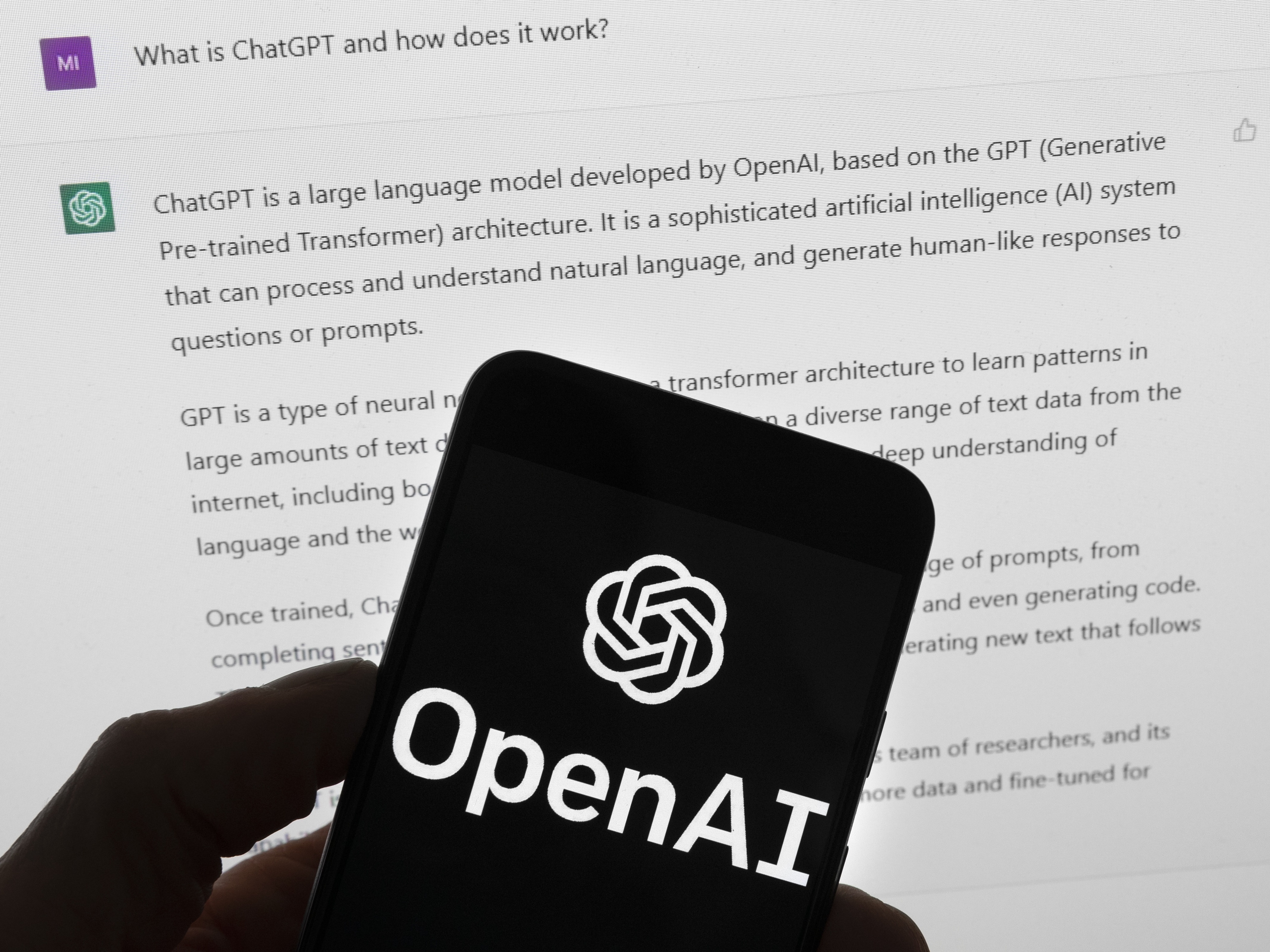 caption: The OpenAI logo is seen on a mobile phone in front of a computer screen which displays output from ChatGPT, March 21, 2023, in Boston.