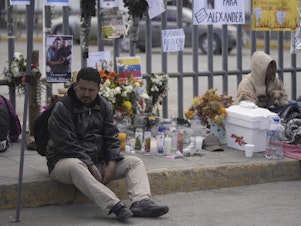 caption: A Venezuelan migrant sits on the sidewalk where an altar was created with candles and photos outside the Mexican immigration processing center that was the site of a deadly fire, as migrants wake up after spending the night on the sidewalk in Ciudad Juárez, Mexico, on Thursday.