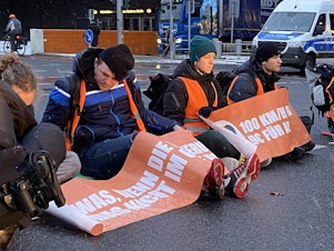 caption: Climate activists from the group Letzte Generation (Last Generation) hold up commuter traffic on a Monday morning in Berlin by supergluing themselves to the road. Police unstick their hands using cooking oil and a pastry brush while irate drivers look on, stuck for more than an hour.