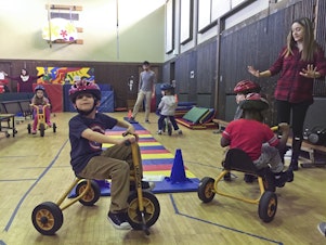 caption: Kindergarteners take turns on the tricycles at the EEU gym class. 
