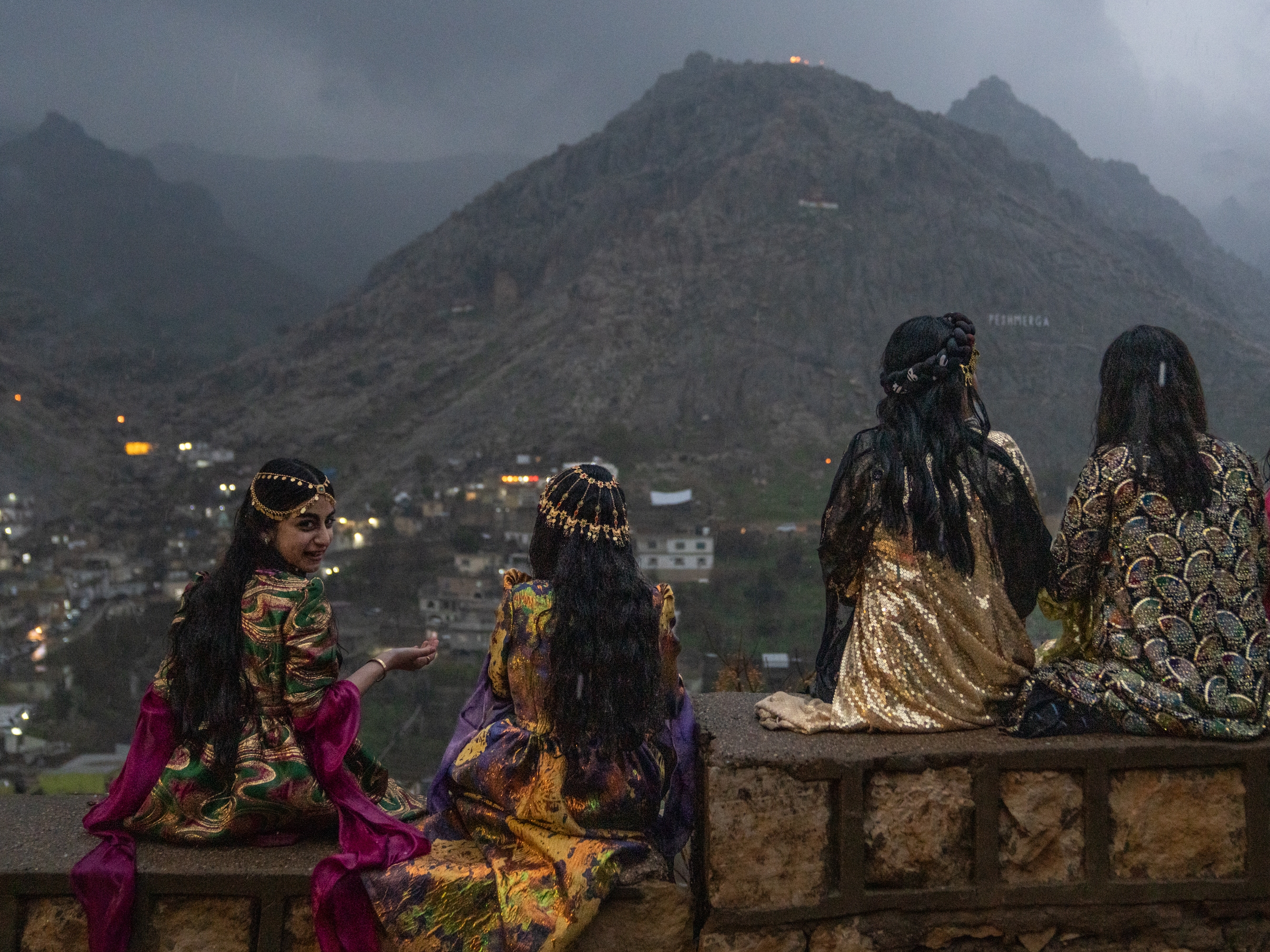 caption: Girls sit on a wall to get a good vantage point of people walking up the mountains with flaming torches and fireworks for Nowruz in Akre, the Kurdish region of Iraq on Friday.
