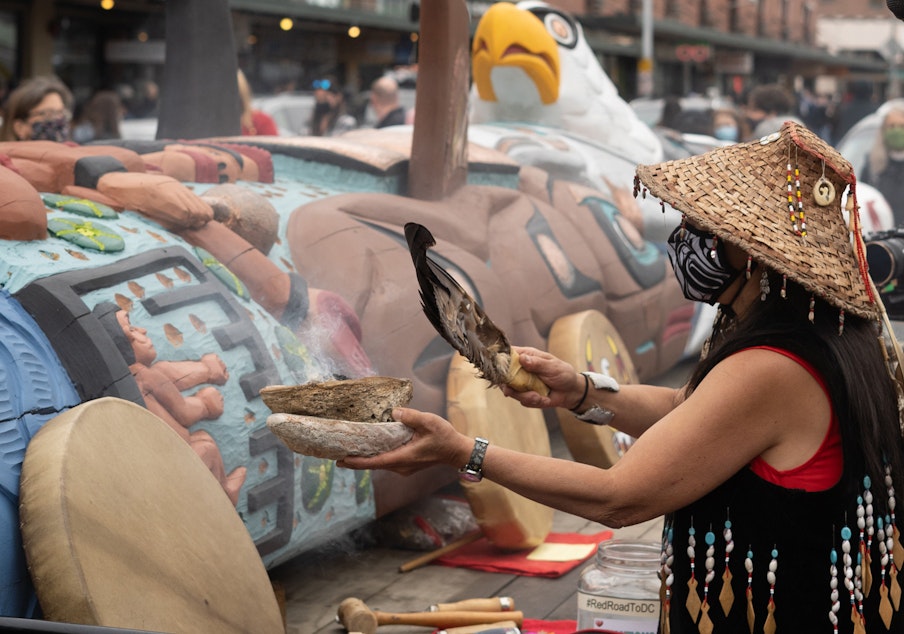 caption: Linda Soriano of the Lummi Nation blesses a 24-foot totem pole, carved by the House of Tears Carvers of the Lummi Nation, with traditional medicine outside of Seattle’s Pike Place Market on Saturday, May 22, 2021, in Seattle. 