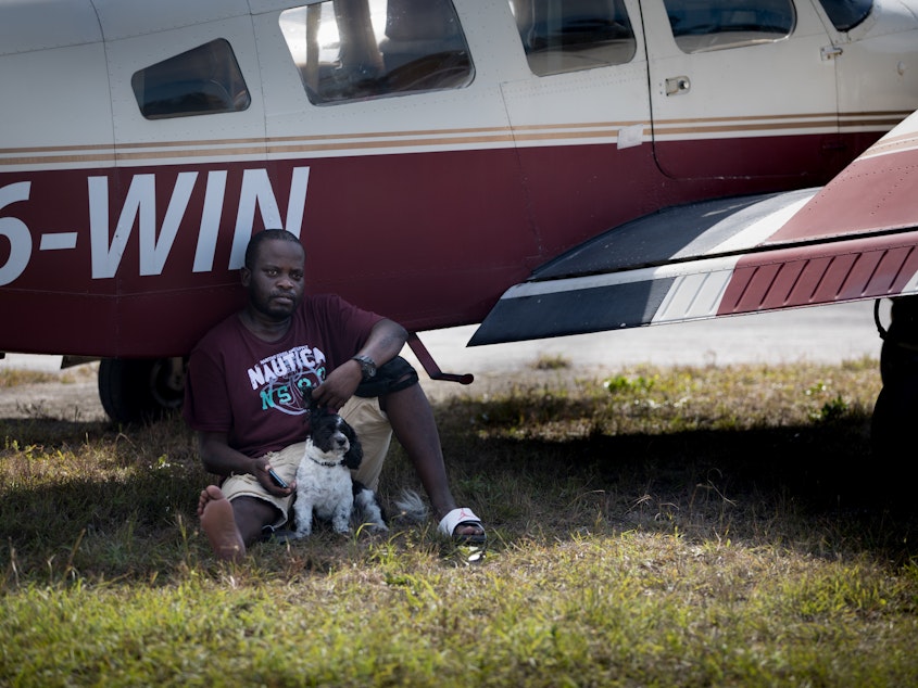 caption: Evacuee Samazio Rolle and his dog, Logan, sit in the shade next to an airplane in the afternoon heat at a landing strip on Abaco, in hopes of catching a flight to Nassau. The pair survived Hurricane Dorian's punishing strike on the Bahamas — and now they want to leave Abaco.