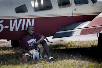 caption: Evacuee Samazio Rolle and his dog, Logan, sit in the shade next to an airplane in the afternoon heat at a landing strip on Abaco, in hopes of catching a flight to Nassau. The pair survived Hurricane Dorian's punishing strike on the Bahamas — and now they want to leave Abaco.