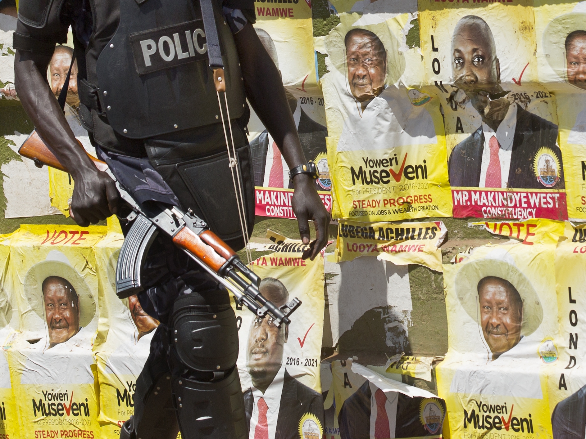 caption: An armed Ugandan riot policeman patrols past campaign posters for longtime President Yoweri Museveni.