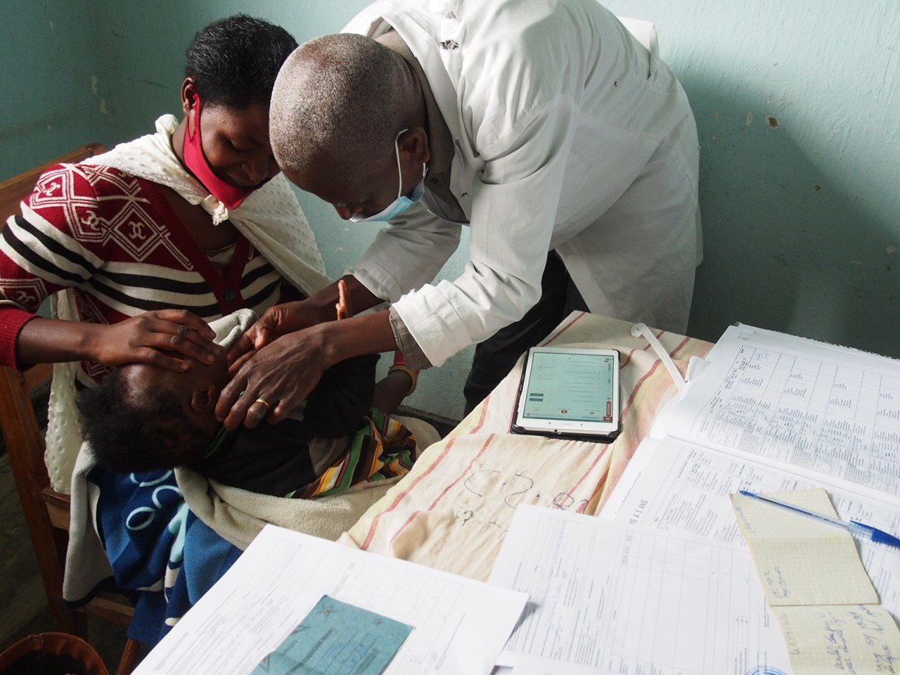 caption: A mother and child at a health center in Rwanda. The child's condition was assessed by a nurse using the new ePOCT+ digital tablet (at the corner of the table), aimed at reducing the overuse of antibiotics.