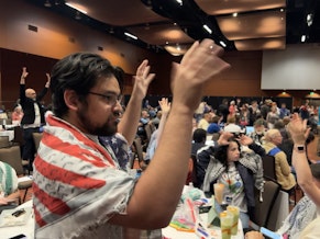 caption: Tariq Yusuf, a delegate to the 2024 Washington State Democratic Convention, raises his hands to show support for passing three resolutions relating to Israel's war in Gaza.