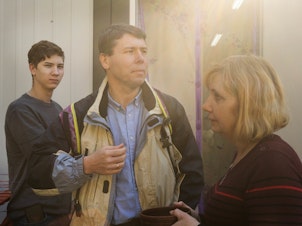 caption: Volodymyr Korchevsky, center, stands with his son Bohdan, 18, and wife, Hanna Korchevska, outside their temporary home in Lviv. The Korchevsky family left the middle-class life they'd built in Mariupol and now months later, jobs gone, savings depleted and unable to afford rent, they are living in what's essentially a short shipping container, sandwiched between others, in a Lviv city park.