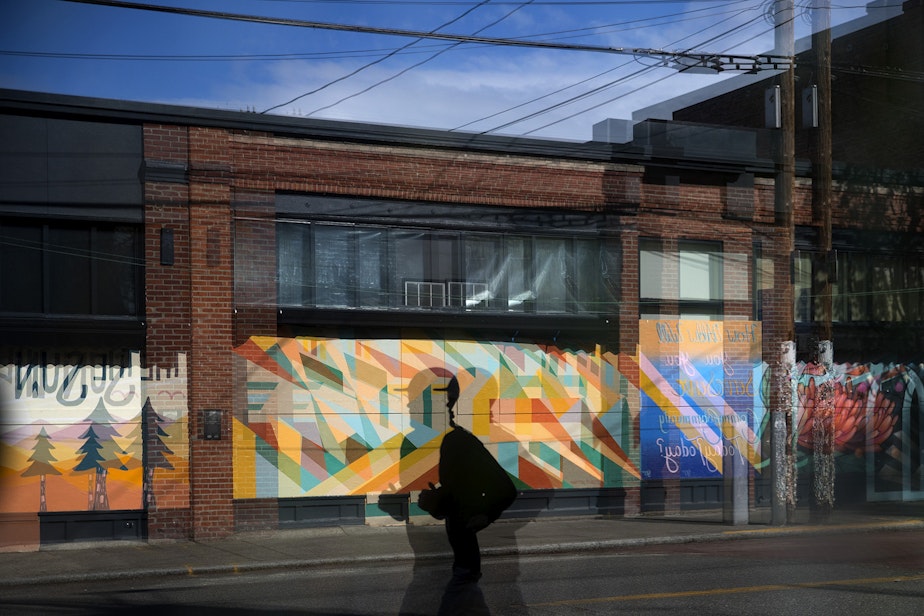 caption: A jogger reflected in the window of a closed business runs along Ballard Avenue in front of painted murals on Sunday, May 3, 2020, near the intersection of 20th Avenue Northwest in Seattle. The murals, from left, were painted by artists Carolyn Wassmer, Sean Mullin and Gynn Rosenberg. 