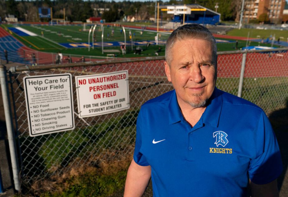 caption: Joe Kennedy, a former assistant football coach at Bremerton High School, poses for a photo March 9, 2022, at the school's football field. He was fired for refusing to stop kneeling in prayer with players and spectators on the field immediately after football games. Kennedy said signs like the one on the fence behind him were put up after he started praying after in efforts to keep spectators from rushing the field to join them. 