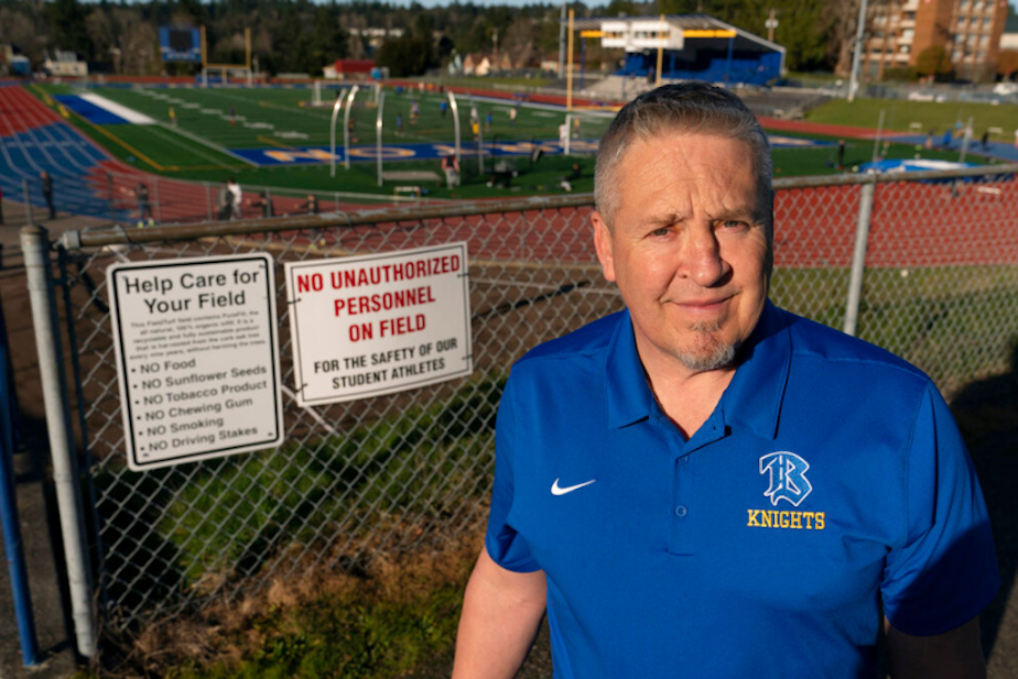 caption: Joe Kennedy, a former assistant football coach at Bremerton High School, poses for a photo March 9, 2022, at the school's football field. He was fired for refusing to stop kneeling in prayer with players and spectators on the field immediately after football games. Kennedy said signs like the one on the fence behind him were put up after he started praying after in efforts to keep spectators from rushing the field to join them. 