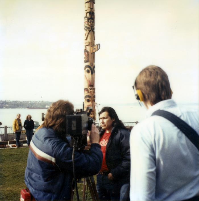 caption: Artist Marvin Oliver (Quinault, Isleta Pueblo) is interviewed by the media in front of one of the totem poles he designed for Victor Steinbrueck Park in 1984.