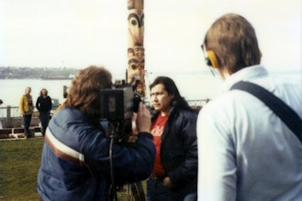 caption: Artist Marvin Oliver (Quinault, Isleta Pueblo) is interviewed by the media in front of one of the totem poles he designed for Victor Steinbrueck Park in 1984.