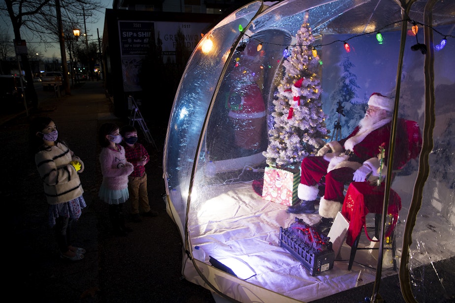 caption: From left, Jasper, 7, Paloma, 6, and Otto, 4, talk with The Seattle Santa, Dan Kemmis, on Sunday, December 6, 2020, as he sits inside of a pandemic safe snow globe at the intersection of Greenwood Avenue North and North 76th Street in Seattle. 