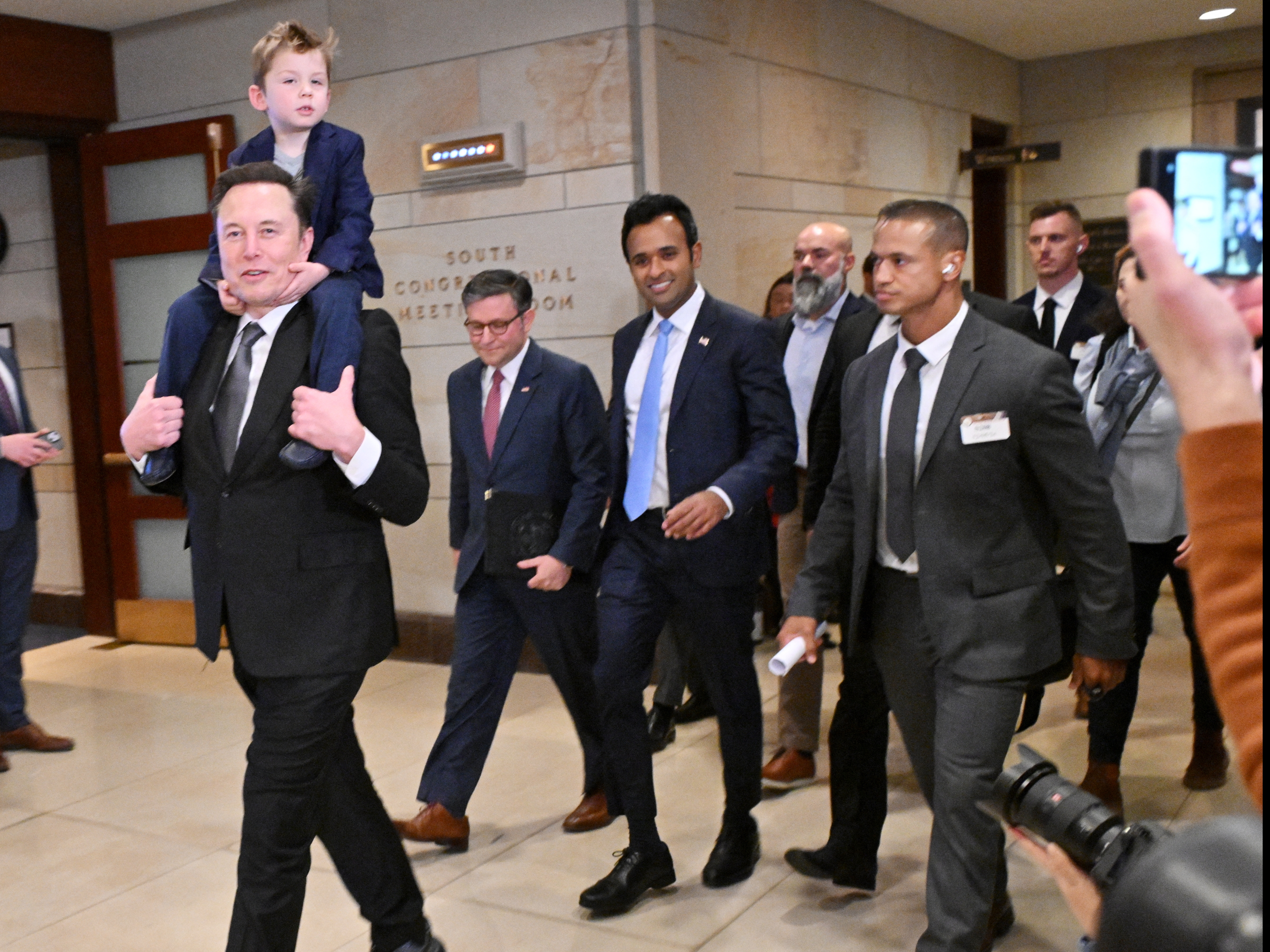 caption: Tesla CEO Elon Musk (left), co-chair of the newly created Department of Government Efficiency (DOGE), carries his son "X" on his shoulders, as he walks with Speaker of the House Mike Johnson (second from left) and DOGE co-leader, businessman Vivek Ramaswamy (center), before a meeting with members of Congress at the Capitol on Dec 5.