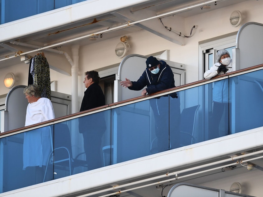 caption: The quarantined Diamond Princess cruise ship has 65 new cases of coronavirus, Japanese officials announced Monday. Here, passengers with ocean-facing rooms stand on their balconies as the ship sits at the Daikoku Pier Cruise Terminal in Yokohama, Japan.