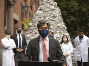 caption: Dr. KJ Seung, a professor at Harvard Medical School, speaks at a September rally outside the home of the CEO of Moderna, maker of one of the two mRNA vaccines that prevent COVID-19. The fake bones are meant to represent lives lost unnecessarily to the coronavirus. Seung was part of a group of doctors demanding Moderna share vaccines — and its recipe — with low-resource countries.