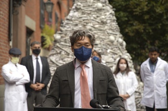 caption: Dr. KJ Seung, a professor at Harvard Medical School, speaks at a September rally outside the home of the CEO of Moderna, maker of one of the two mRNA vaccines that prevent COVID-19. The fake bones are meant to represent lives lost unnecessarily to the coronavirus. Seung was part of a group of doctors demanding Moderna share vaccines — and its recipe — with low-resource countries.
