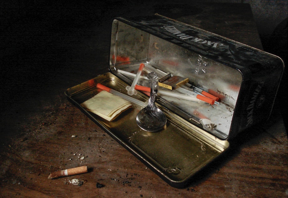 On a dark wooden surface, a black metal box is tipped onto its side, with a spoon and syringes tumbling out over the lid.