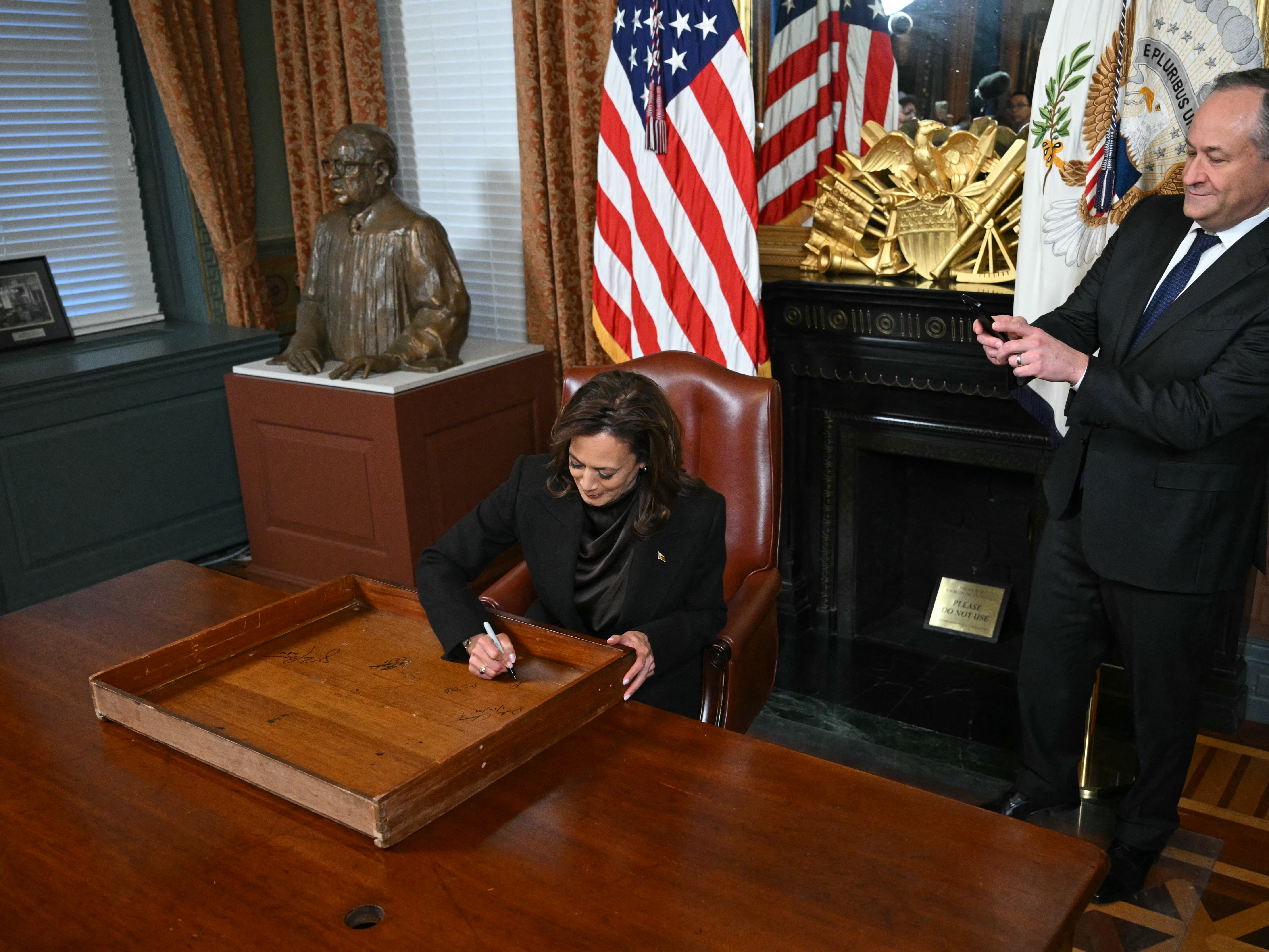 caption: Vice President Harris signs the drawer of her desk as second gentleman Doug Emhoff takes a picture on Jan. 16, 2025.